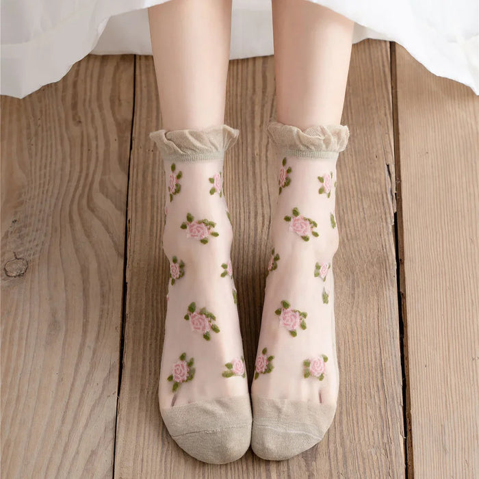 A pair of feet in Lacatang Shop's Kawaii Harajuku style Vintage Floral Lace Ruffle Socks for women, featuring pink rose patterns, a frilled top edge, and solid toe area, stand on a wooden floor, showcasing their sheer yet charming allure.