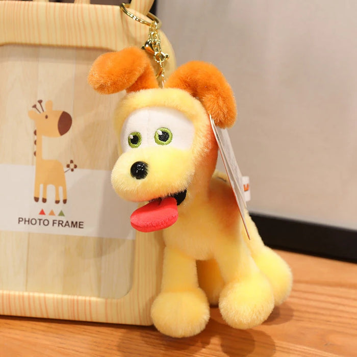 A Kawaii Oudi Dog plush keychain from Lacatang Shop, featuring a smiling yellow face and red tongue, hangs on a gold chain. It sits on wood next to a giraffe-themed frame, adding charm to your plush collection. Perfect for room décor or as an adorable gift for kids and girls.