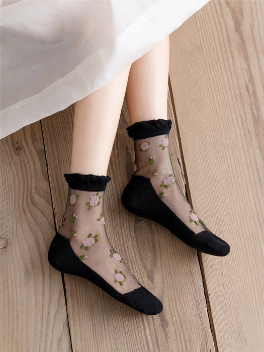 The individual wears Lacatang Shop's Delicate Vintage Floral Lace Ruffle Socks, featuring sheer black fabric adorned with pink and green lace embroidery. These kawaii Harajuku style socks have black toe, heel, and cuff details, beautifully complementing the wooden floor setting partially draped in white.