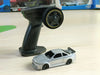 The Lacatang Shop's 2.4G RC Drift Car 1/43 4WD Remote Control Car, a sleek silver model complete with a rear spoiler, is positioned next to its black and gray steering wheel-style remote control on a light wooden surface. Several toy boxes and cars are blurred in the background.