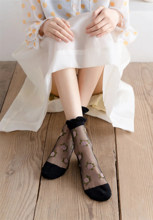 A person sits cross-legged on a wooden floor in a white dress with translucent polka dot sleeves and wears Lacatang Shop's Delicate Vintage Floral Lace Ruffle Socks with Crystal Silk, featuring sheer black fabric and black toes—epitomizing kawaii Harajuku style.