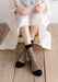 A person sits cross-legged on a wooden floor in a white dress with translucent polka dot sleeves and wears Lacatang Shop's Delicate Vintage Floral Lace Ruffle Socks with Crystal Silk, featuring sheer black fabric and black toes—epitomizing kawaii Harajuku style.