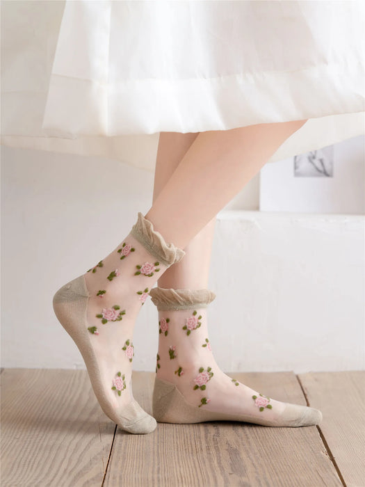 A person wears Lacatang Shop's Vintage Floral Lace Ruffle Socks for Women, Kawaii Harajuku style ultra-thin transparent crystal silk crew socks with pink rose patterns. They pair them with a white skirt against neutral-toned walls and wooden flooring, creating a serene backdrop.