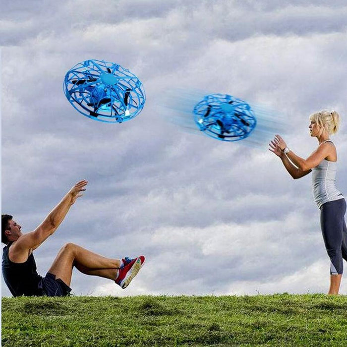 A man sitting on grass leans back with his hands raised as two blue Jade Flying Helicopter Mini drones, among the best products in electric toys, hover above him. A woman in athletic attire stands nearby with her arms extended towards the drones. The sky is cloudy in the background.