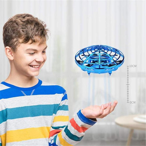 A smiling boy with curly hair wearing a colorful striped sweater holds his hand up, while the Jade Flying Helicopter Mini Drone UFO RC Drone Infrared Induction Aircraft hovers above it indoors. The background features light-colored curtains, and a scale indicating height from 0 to 10 cm is visible.